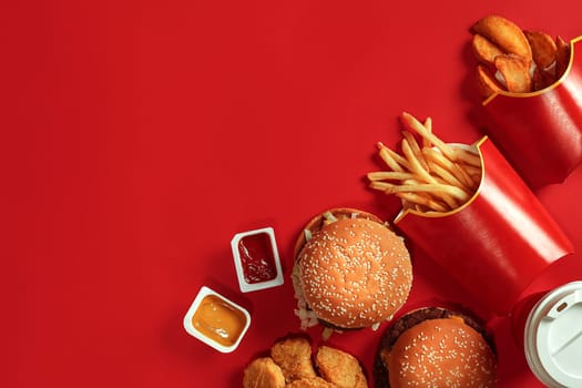 Fast food dish top view. Meat burger, potato chips and glass of drink on red background. Takeaway composition. Wrapped French fries, hamburger, mayonnaise and ketchup sauces on red desk. Copy space