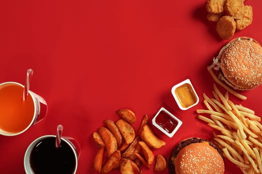 Fast food dish top view. Meat burger, potato chips and glass of drink on red background. Takeaway composition. Wrapped French fries, hamburger, mayonnaise and ketchup sauces on red desk. Copy space