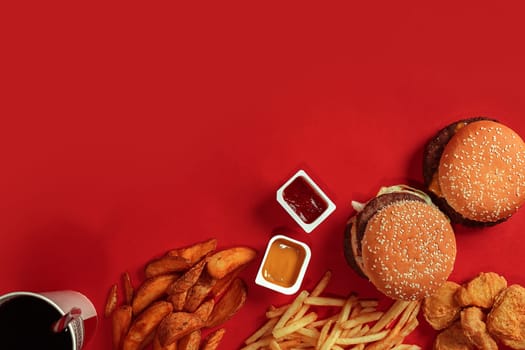 Fast food dish top view. Meat burger, potato chips and glass of drink on red background. Takeaway composition. Wrapped French fries, hamburger, mayonnaise and ketchup sauces on red desk. Copy space
