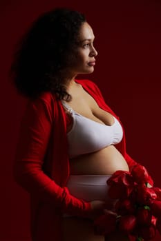 Fashion portrait of a beautiful multi ethnic pregnant woman, wearing white underwear and red unbuttoned shirt, holding a bunch of red tulips, posing bare belly, standing side to a dark red background