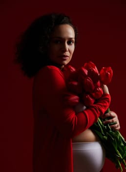 Authentic portrait of adult pregnant woman holding a bouquet of tulips, looking confidently at camera, isolated on dark background. Pregnancy fashion. Maternity concept. Happy Mother's, Women's Day