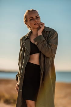 Portrait blonde sea cape. A calm young blonde in an unbuttoned khaki raincoat stands on the seashore, under the raincoat there is a black skirt and top.