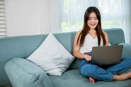 Happy woman typing email on notebook computer, Asian young female smiling sitting relaxing on sofa using laptop in living room at home, freelance browsing through the internet during free time