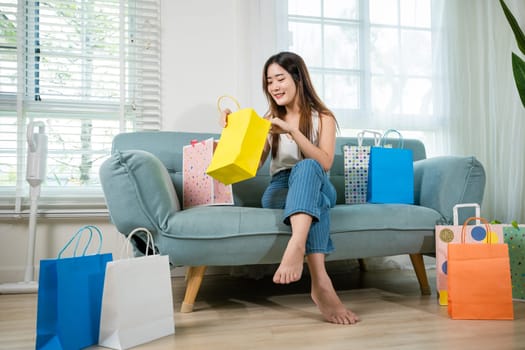 Happy Asian young woman with many shopping bags at home in living room after long day shopping, Excited female online shopping sitting on couch, Shopper or shopaholic concept