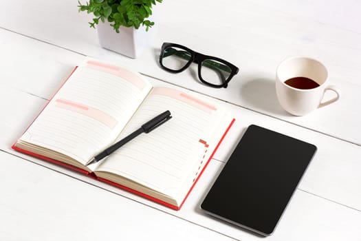 Office table desk with set of supplies, white blank notepad, cup, pen, tablet, glasses, flower on white background. Top view and copy space for text