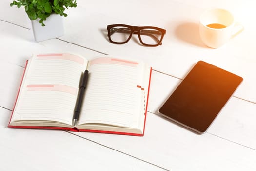 Office table desk with set of supplies, white blank notepad, cup, pen, tablet, glasses, flower on white background. Top view and copy space for text. sun flare