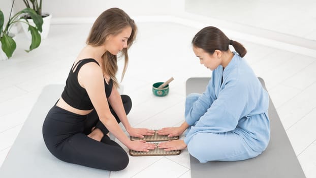 Two women sit on yoga mats with their hands on sadhu boards