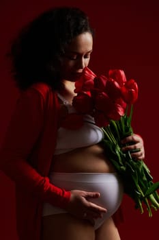 Side portrait of multi ethnic pregnant woman, expectant mother sniffing a bouquet of red tulips, gently stroking her belly, isolated on dark red background. Femininity, sensuality, pregnancy fashion