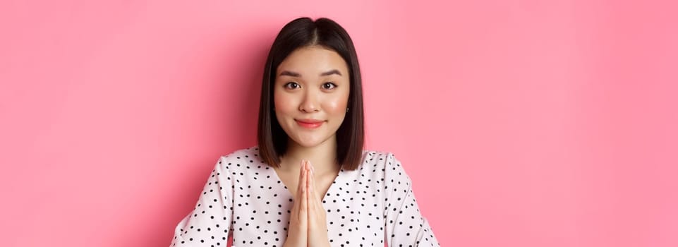 Beauty and lifestyle concept. Close-up of cute asian woman smiling, showing thank you gesture, holding hands in pray, standing over pink background.