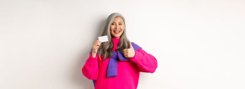 Shopping concept. Smiling asian middle-aged woman with grey hair showing plastic credit card and thumbs-up, recommending bank promotion, white background.