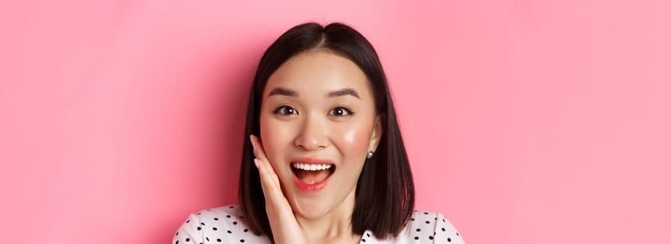 Beauty and lifestyle concept. Close-up of surprised and happy asian woman staring at camera amazed, gasping with disbelief, standing over pink background.