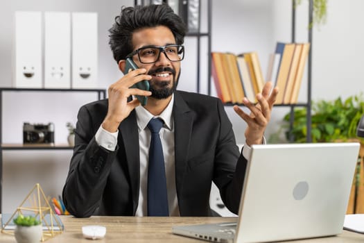 Bearded indian businessman working at office, having mobile phone talk at office desk with laptop. Manager freelance indian man holding smartphone using messenger chat apps. Employment, occupation