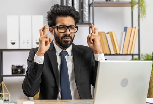 Bearded indian businessman crossed fingers looking at laptop screen asking for good luck news, wishing good exam results, dreaming about win victory in lottery jackpot, business money profit at office