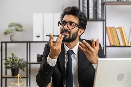 Bearded indian businessman working, having mobile phone loudspeaker talk at office desk with laptop. Manager freelance indian man holding smartphone using messenger chat apps. Employment, occupation