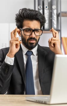 Bearded indian businessman crossed fingers looking at laptop screen asking for good luck news, wishing good exam results, dreaming about win victory in lottery jackpot, business money profit at office