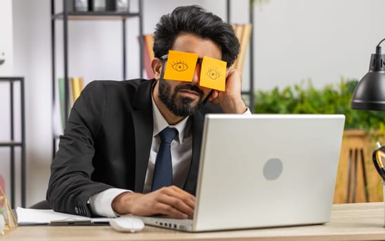 Inefficient tired young businessman working sleeping on laptop computer with eyes stickers on face at office workplace desk. Indian lazy manager freelancer man. Business people cheating to sleep