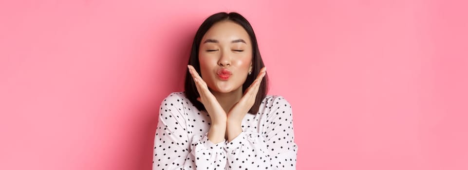 Beauty and skin care concept. Close-up of beautiful asian girl close eyes, pucker lips for kisses, showing cute perfect face, standing over pink background.