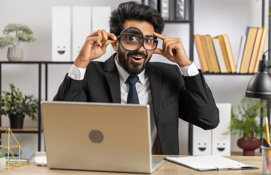 Investigator researcher scientist businessman working at home office holding magnifying glass near face looking into camera with big zoomed funny eye, searching, analysing. Professional freelancer man