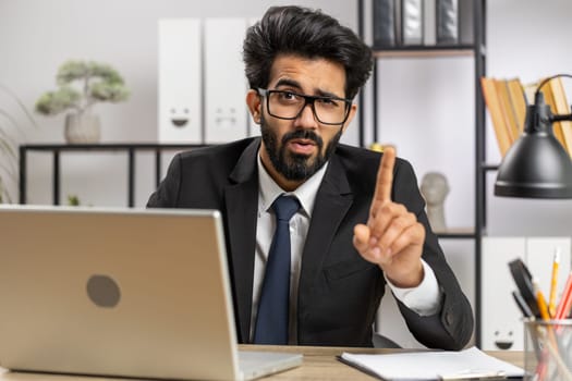 Indian young businessman working on laptop computer shakes finger and saying No be careful scolding and giving advice to avoid danger mistake disapproval sign at home office. Confident freelancer man