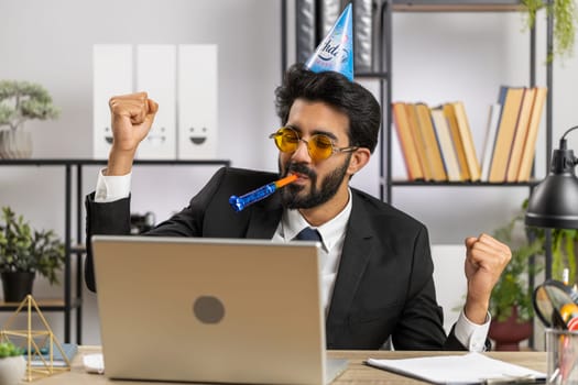 Excited lucky business man in sunglasses, festive cap hat cone blow pipe at workplace dancing victory dance, weekend. Rejoicing developing project. Celebrating birthday. Lottery jackpot. Office people