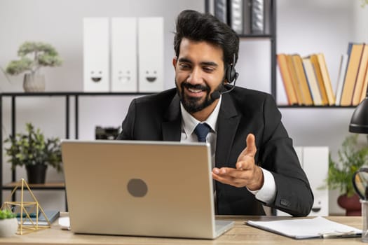 Indian business man working on laptop wearing headset, freelance worker call center or support service operator helpline, having pleasant talk with client or colleague communication support at office