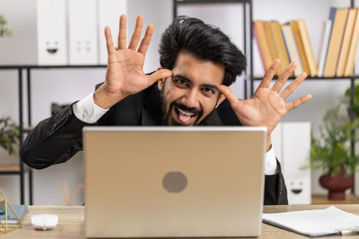 Indian businessman hiding behind laptop computer, making funny face, fooling around, putting his tongue out remote working disrespecting someone at office desk workplace. Professional freelancer man