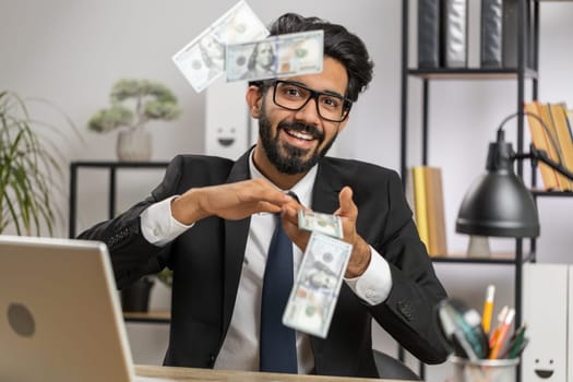 Cheerful rich young indian businessman working on laptop and wasting throwing money to camera more tips earnings big profit success win lottery shopping. Happy freelancer arab man at home office desk
