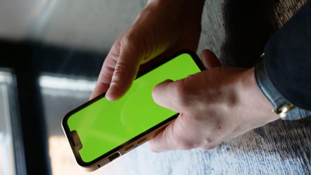 Close-up smartphone with green screen in men's hands. A businessman scrolls the phone with chroma key in the office. Vertical