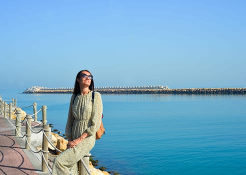 Young happy woman in a cute dress sits on a pier by the sea enjoying summer sunbeams.