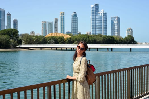 The skyline of Sharjah is visible from the waterfront - a woman tourist with a backpack enjoys the view of the water and skyscrapers. Cityscape of Sharjah UAE