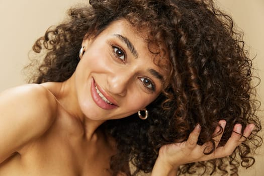 Woman applies cream and balm to her curly hair, the concept of protection and care, a healthy look, a smile with teeth on a beige background.