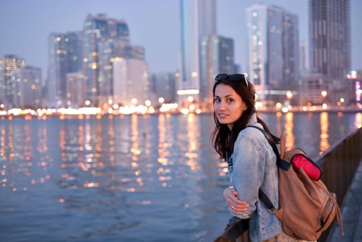 A tourist girl with a backpack on her shoulders enjoys a view of the modern skyscrapers of the Sharjah marina at night.