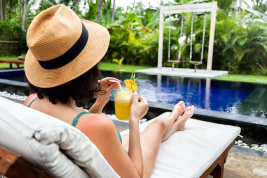 Woman relaxing sitting in desk chair near swimming pool drinking healthy pineapple juice during summer holidays. Copy space. Vacation concept.
