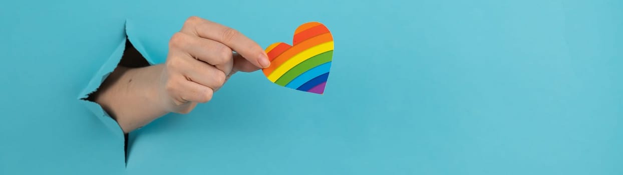 A hand with a rainbow-colored heart sticking out of a hole in a blue cardboard background. Widescreen
