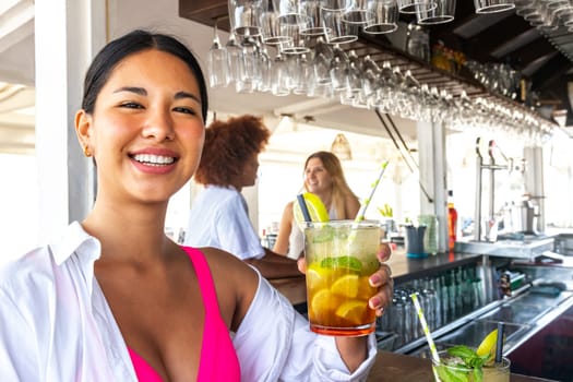 Happy young latina woman showing mojito cocktail looking at camera at a beach bar in summertime. Vacation concept.