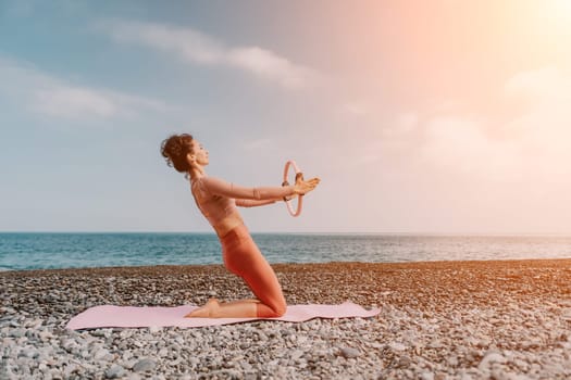 Middle aged well looking woman with black hair doing Pilates with the ring on the yoga mat near the sea on the pebble beach. Female fitness yoga concept. Healthy lifestyle, harmony and meditation.