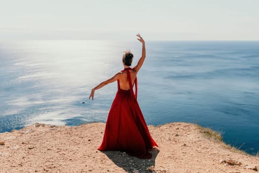 Side view a Young beautiful sensual woman in a red long dress posing on a rock high above the sea during sunrise. Girl on the nature on blue sky background. Fashion photo.