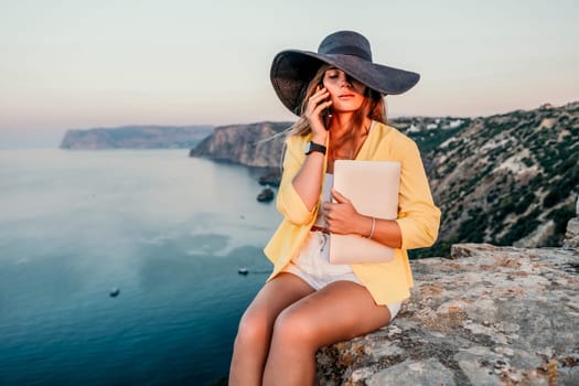 Successful business woman in yellow hat working on laptop by the sea. Pretty lady typing on computer at summer day outdoors. Freelance, travel and holidays concept.