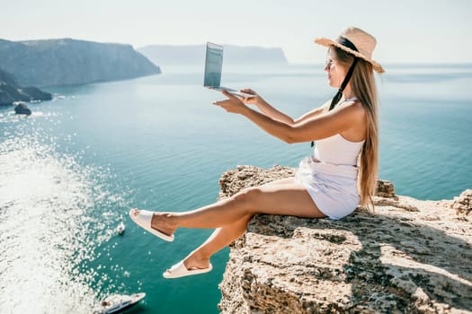 Working remotely on seashore. Happy successful woman female freelancer in straw hat working on laptop by the sea at sunset. Freelance, remote work on vacation