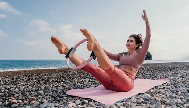 Middle aged well looking woman with black hair doing Pilates with the ring on the yoga mat near the sea on the pebble beach. Female fitness yoga concept. Healthy lifestyle, harmony and meditation.