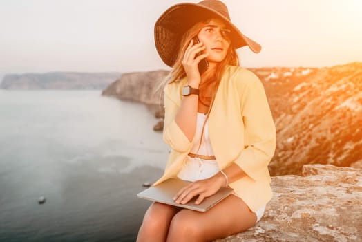 Successful business woman in yellow hat working on laptop by the sea. Pretty lady typing on computer at summer day outdoors. Freelance, travel and holidays concept.