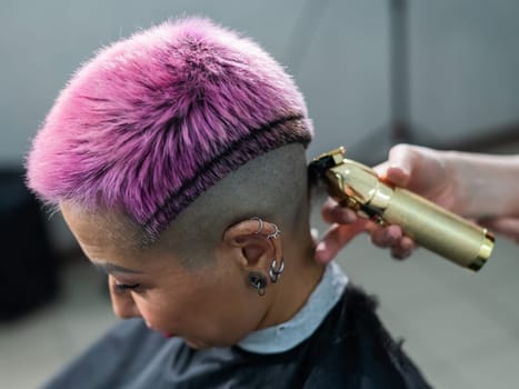 The hairdresser shaves the temple of a female client. Asian woman with short pink hair in barbershop