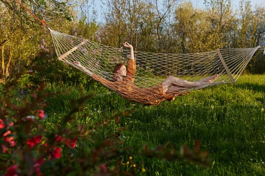 a happy woman in a long orange dress is relaxing in nature lying in a mesh hammock enjoying summer and vacation in the country surrounded by green foliage, happily lifting her legs. High quality photo