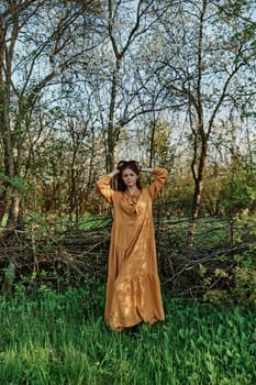 a beautiful woman walks in the shade near the trees, dressed in a long orange dress, enjoying the weather and the weekend, straightening her hair with her hands. The theme of privacy with nature,. High quality photo