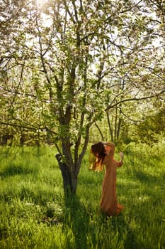 a beautiful, joyful woman stands in a long orange dress, in the countryside, near a tree blooming with white flowers, during sunset, illuminated from behind and touches her long hair. High quality photo