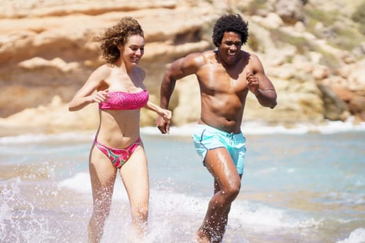 Cheerful multiracial couple in swimwear running in splashing seawater and looking down while enjoying summertime together near seaside against blurred rocks