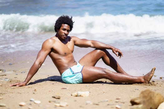 Full body side view of African American male relaxing on sandy beach with arm on knee while looking at camera, and leaning on hand near waving foamy sea