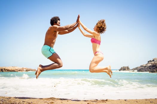 Full length side view of cheerful African American man and woman in bikini jumping on sandy seashore and holding hands while looking at each other