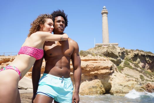 From below of positive multiracial boyfriend and girlfriend in swimwear standing on seashore and embracing each other against lighthouse on hill while looking away