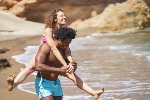 Cheerful young African American man giving piggyback ride to curly haired woman in bikini having fun on sandy beach near waving sea